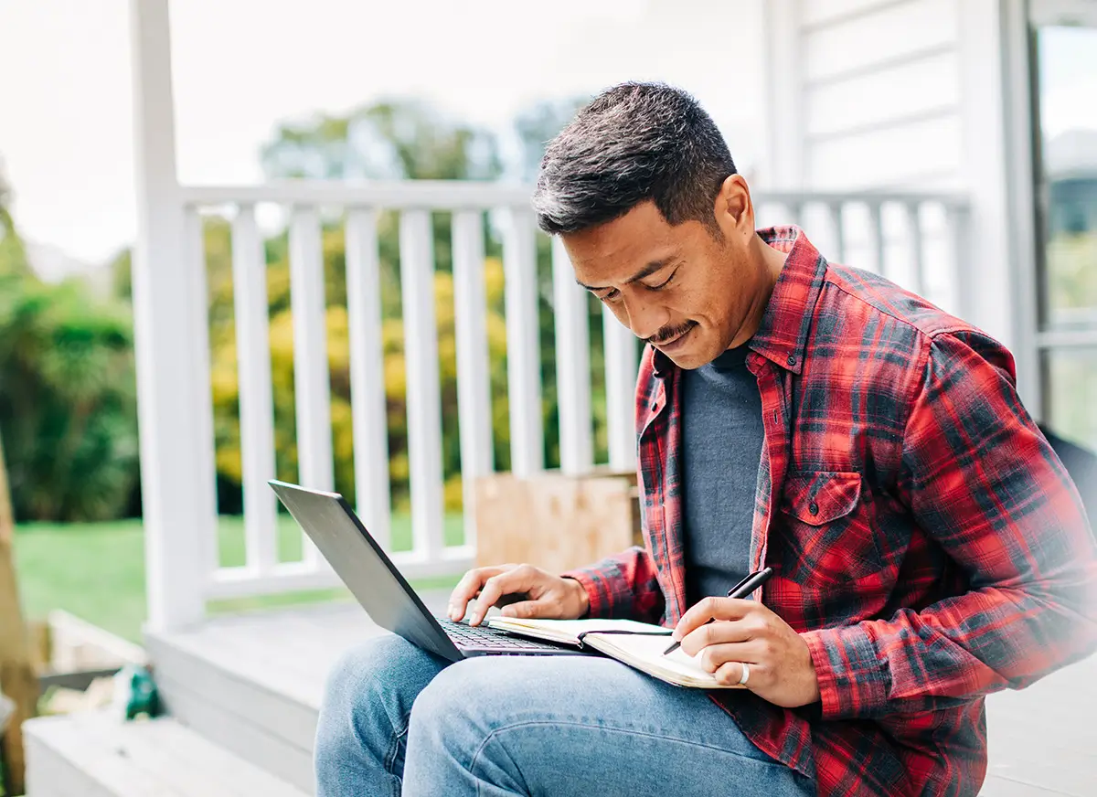 Polynesian man sitting outside a house working on laptop and writing notes