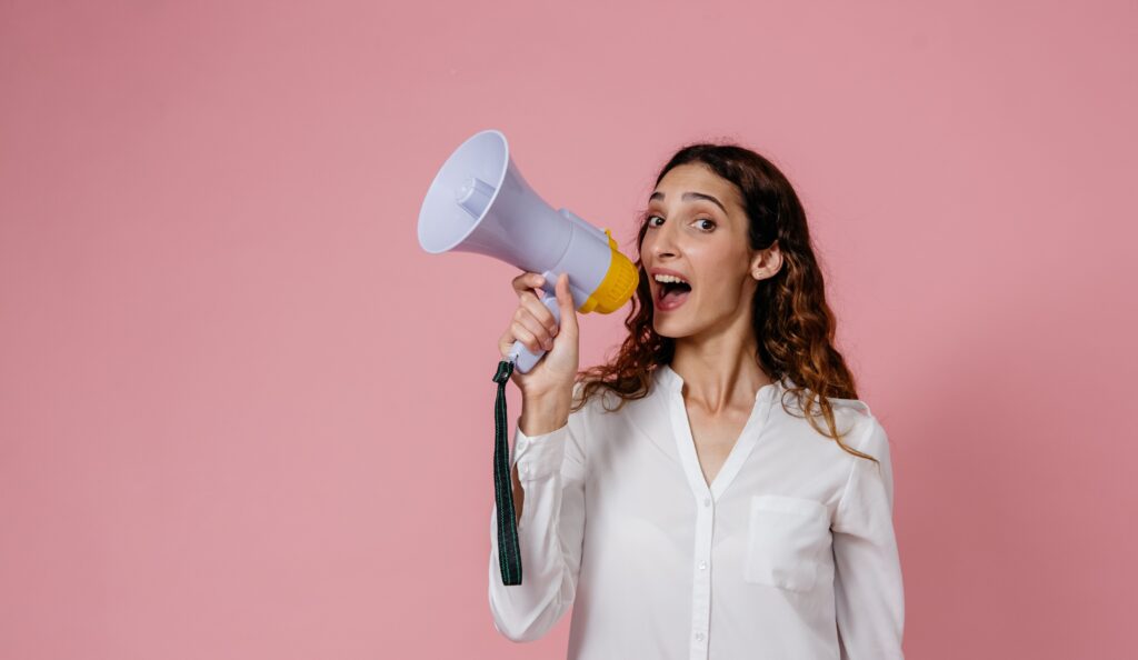 Woman with curly hair shouts into a megaphone against a pink background